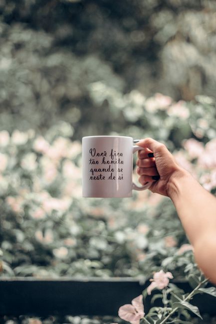 A close-up of a person's hand holding a white ceramic mug with black cursive writing, set against a blurred garden background with flowering plants and leafy trees. The garden features well-maintained